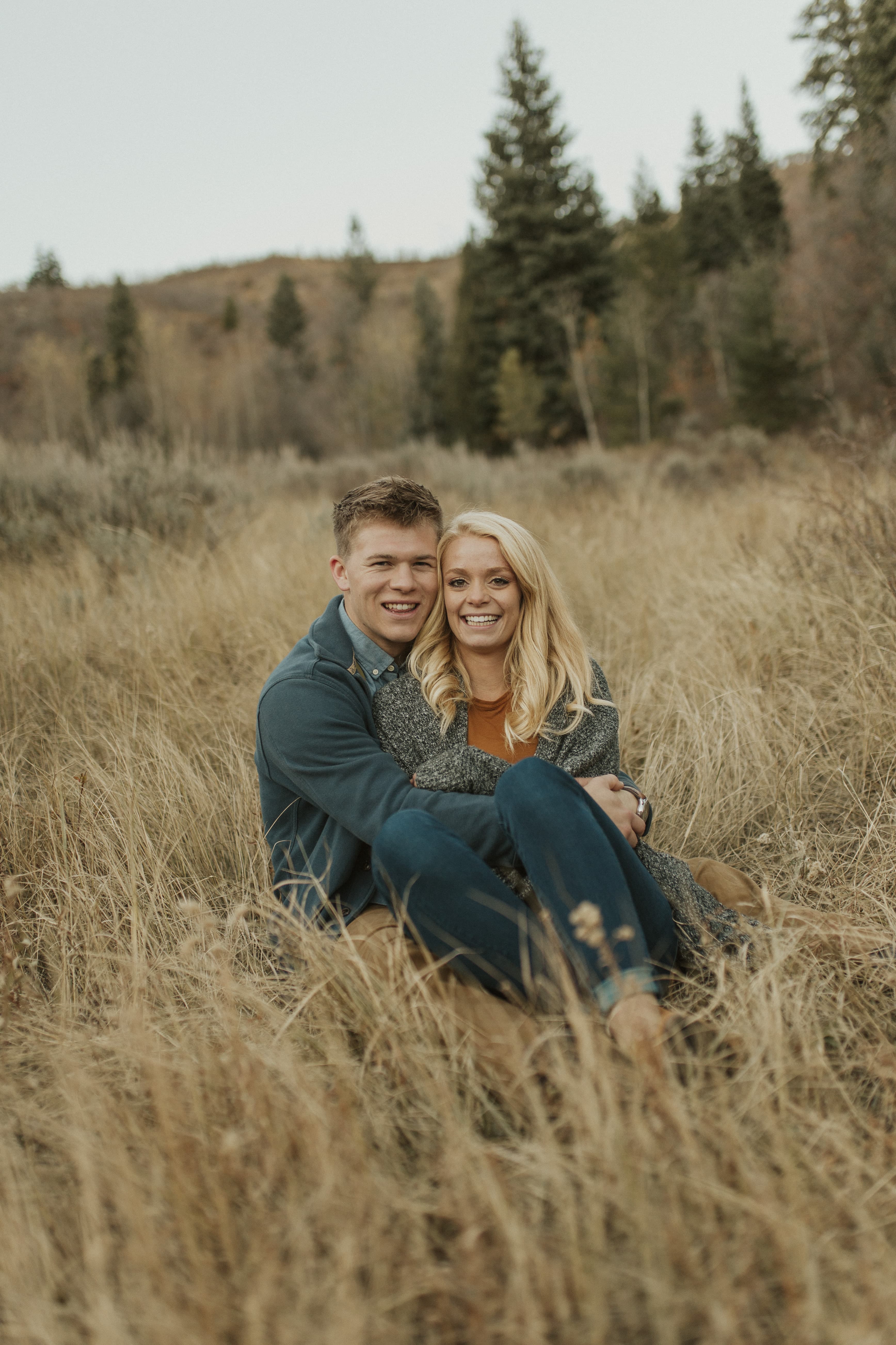 A smiling young couple sits together in a field of tall, dry autumn grass.