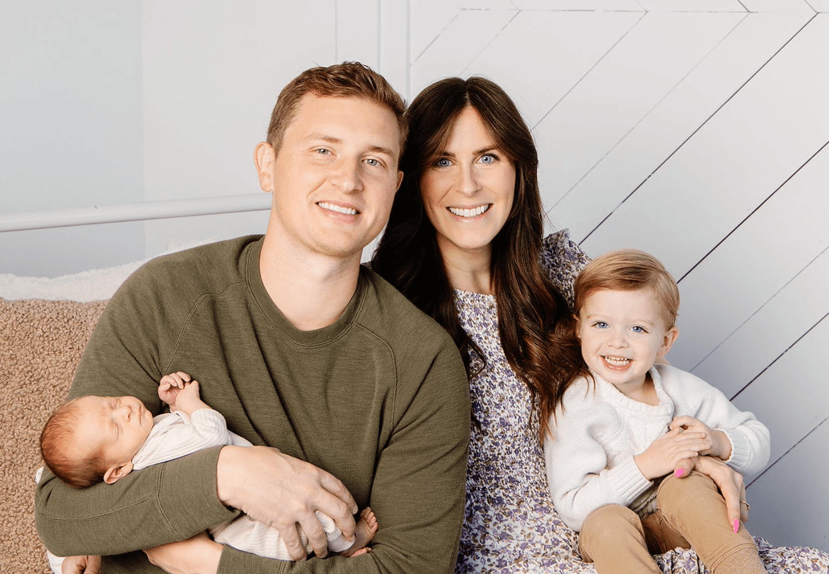 Smiling parents pose with their toddler and sleeping newborn baby against a white wall.